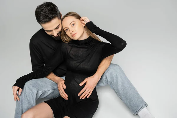 Esposo en camiseta negra y jeans tocando el vientre de la esposa embarazada de moda en vestido mirando a la cámara mientras está sentado en un fondo gris, nuevos comienzos y concepto de crianza - foto de stock