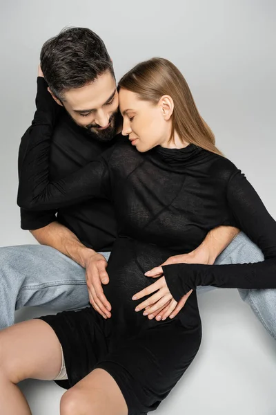 Mujer de pelo justo de moda y embarazada en vestido negro abrazando al marido barbudo mientras están sentados juntos sobre un fondo gris, nuevos comienzos y concepto de crianza - foto de stock