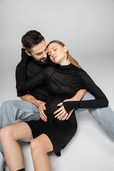 Smiling and bearded man in t-shirt and jeans hugging fashionable and pregnant wife while sitting together on grey background, new beginnings and parenting concept — Stock Photo