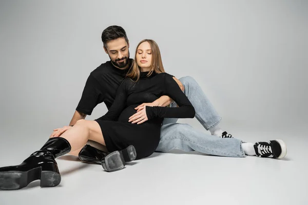 Bearded man in jeans and t-shirt touching belly of fair haired and stylish pregnant wife in black dress while sitting on grey background, new beginnings and parenting concept — Stock Photo