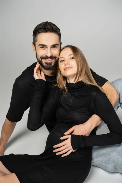 Homem barbudo alegre em camiseta preta abraçando a mulher grávida na moda no vestido e olhando para a câmera enquanto sentado em fundo cinza, novos começos e conceito de parentalidade — Fotografia de Stock