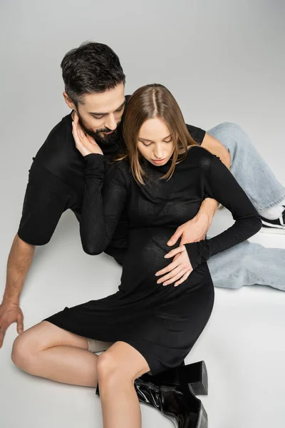 High angle view of fashionable pregnant woman in black dress touching husband and looking at belly while sitting on grey background, new beginnings and parenting concept — Stock Photo