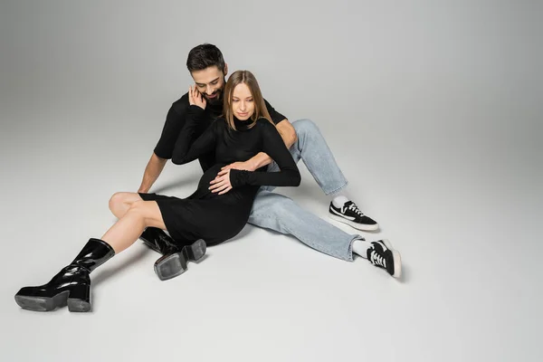 Smiling bearded man in jeans and t-shirt hugging fashionable pregnant woman in dress while sitting on grey background, new beginnings and parenting concept, husband and wife — Stock Photo