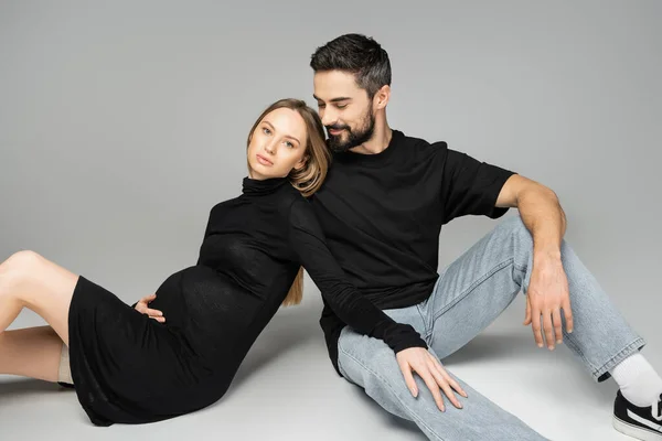 Позитивний бородатий чоловік у чорній футболці та джинсах дивиться на модну та вагітну дружину в одязі, сидячи разом на сірому фоні, нові початку та концепцію виховання — стокове фото