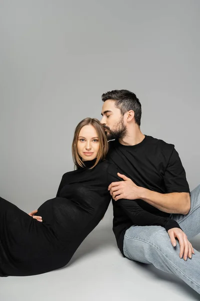 Бородатий чоловік в джинсах і чорній футболці цілує модну вагітну жінку в одязі і сидить на сірому фоні, нові починання і концепція батьківства, чоловік і дружина — Stock Photo