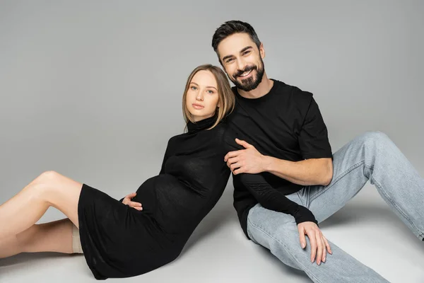 Cheerful husband in t-shirt and jeans looking at camera and hugging trendy pregnant woman in dress while sitting on grey background, new beginnings and parenting concept, husband and wife — Stock Photo