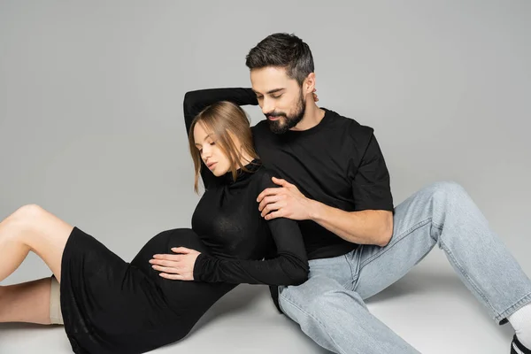Homem barbudo em jeans e camiseta preta abraçando mulher elegante e grávida em vestido preto enquanto sentado em fundo cinza, novos começos e conceito de parentalidade, marido e mulher — Fotografia de Stock