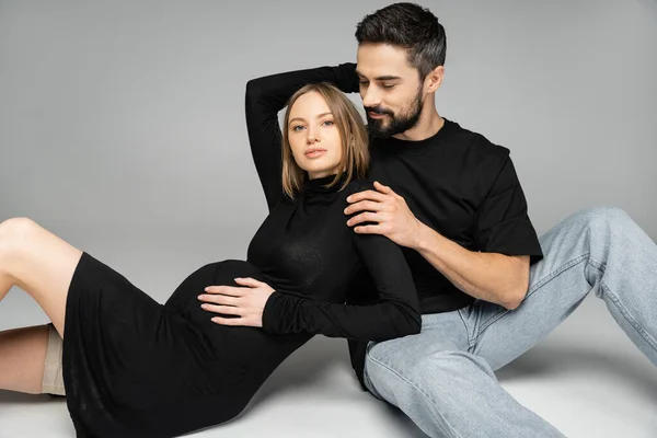 Stylish pregnant woman in black dress hugging bearded man in t-shirt and jeans while looking at camera on grey background, new beginnings and parenting concept, husband and wife — Stock Photo