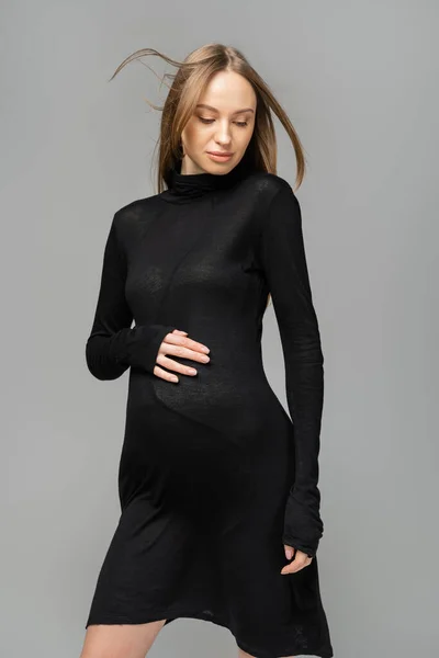 Модная и светловолосая беременная женщина с натуральным макияжем, позирующим в черном платье и трогательным животом, изолированным от серого, новое начало и концепция материнства, будущая мать — стоковое фото
