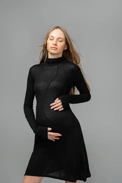 Relajada mujer cabellera y embarazada en elegante vestido negro cerrando los ojos y tocando el vientre mientras está de pie aislado en gris, nuevos comienzos y el concepto de maternidad, madre-a-ser - foto de stock