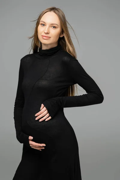 Portrait of hair haired and pregnant woman with natural makeup wearing black dress and looking at camera while standing isolated on grey, new beginnings and maternity concept, mom-to-be — Stock Photo