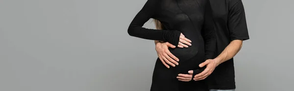 Обрезанный вид мужчины в черной футболке, обнимающего стильную беременную жену в платье, стоя вместе изолированно на сером с копировальным пространством, новыми начинаниями и концепцией предвкушения, баннером — стоковое фото