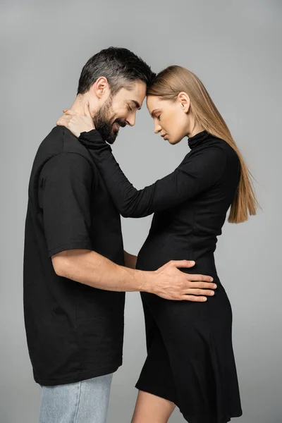 Side view of stylish and pregnant fair haired woman in black dress hugging smiling and bearded husband while standing with closed eyes isolated on grey, new beginnings and anticipation concept — Stock Photo