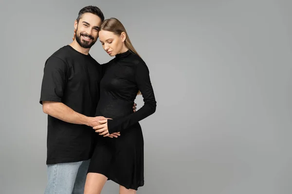 Joyful and bearded man in black t-shirt and jeans hugging pregnant wife in dress and looking at camera while standing together isolated on grey, new beginnings and anticipation concept — Stock Photo