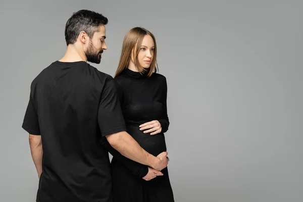 Hombre barbudo en camiseta negra tocando el vientre de la esposa de moda y embarazada en el vestido, mientras que de pie juntos aislados en gris con espacio de copia, nuevos comienzos y concepto de anticipación - foto de stock