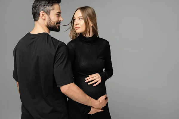 Stylish and pregnant woman in black dress touching belly and looking at bearded husband in t-shirt while standing isolated on grey, new beginnings and anticipation concept — Stock Photo