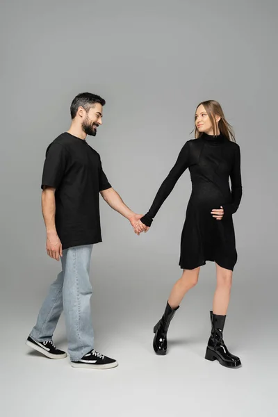 Полная длина стильной и беременной женщины в черном платье, держащей за руку улыбающегося мужа и идущей вместе на сером фоне, новые начинания и концепция ожидания — стоковое фото