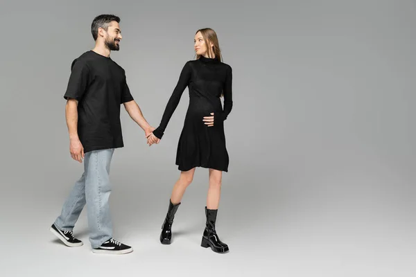 Full length of fashionable pregnant woman in dress holding hand of cheerful husband in jeans and black t-shirt while walking on grey background, new beginnings and anticipation concept — Stock Photo