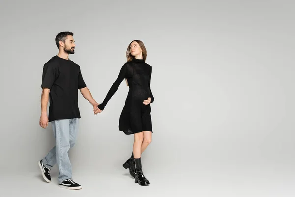 Full length of trendy pregnant woman in dress walking and holding hand of bearded husband in t-shirt and jeans on grey background, new beginnings and anticipation concept, expecting parents — Stock Photo