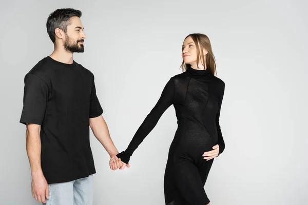 Smiling bearded man in black t-shirt holding hand of fair haired and pregnant wife in stylish dress on grey background, new beginnings and anticipation concept, expecting parents — Stock Photo