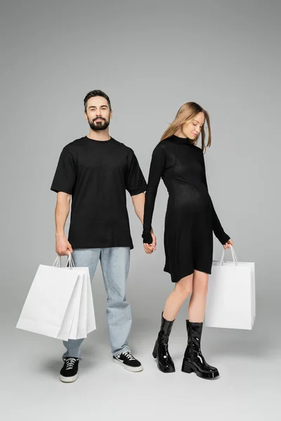 Full length of stylish and pregnant woman in black dress holding shopping bags and hand of husband while standing on grey background, new beginnings and parenthood concept — Stock Photo