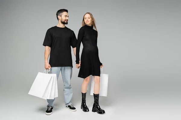 Усміхнений бородатий чоловік у футболці і джинсах тримає сумки для покупок і руку стильної вагітної дружини і стоїть разом на сірому фоні, нові початку і концепція батьківства — стокове фото