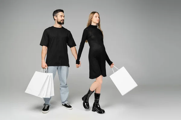 Fashionable pregnant woman in black dress holding shopping bags and hand of husband while walking on grey background, new beginnings and parenthood concept — Stock Photo
