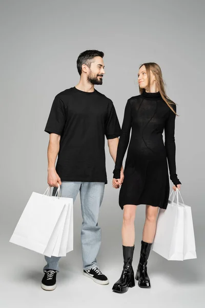 Hombre barbudo sonriente sosteniendo bolsas de compras y mirando a la esposa embarazada de moda en vestido y de pie sobre fondo gris, nuevos comienzos y concepto de paternidad - foto de stock