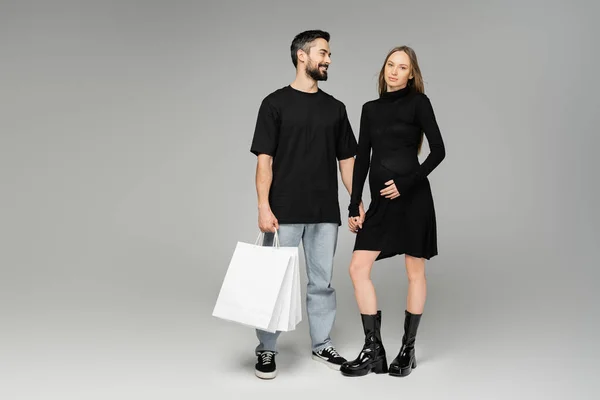Joyful bearded man holding shopping bags and looking at trendy pregnant wife in black dress on grey background, new beginnings and parenthood, shopping and expectation concept — Stock Photo