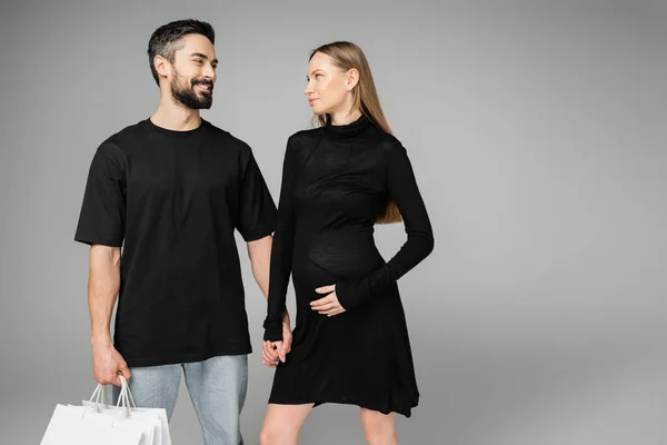 Модная беременная женщина в черном платье держит за руку улыбающегося мужа с пакетами для покупок, стоя вместе изолированные на сером, новых начинаниях и концепции родительства — стоковое фото