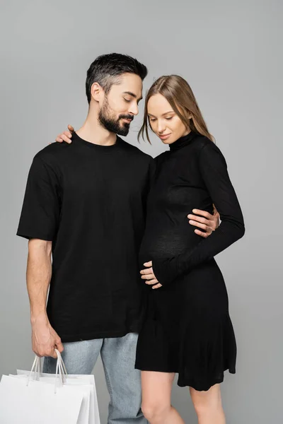 Bearded man in jeans and t-shirt holding shopping bags and hugging pregnant wife in stylish black dress while standing isolated on grey, new beginnings and parenthood concept — Stock Photo