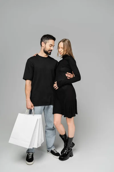 Man in jeans and t-shirt holding shopping bags and hugging trendy pregnant wife in black dress and standing together on grey background, new beginnings and shopping concept — Stock Photo