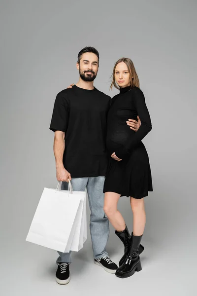 Comprimento total da mulher grávida na moda em vestido preto abraçando marido sorridente com sacos de compras e olhando para a câmera em fundo cinza, novos começos e conceito de compras — Fotografia de Stock