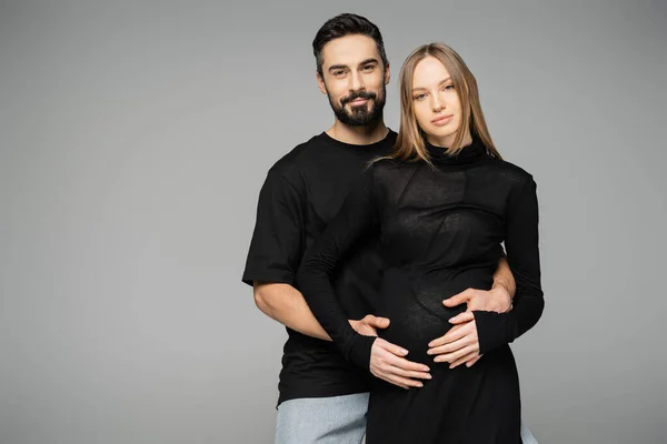 Бородатый и улыбающийся мужчина в черной футболке, обнимающий стильную беременную жену в платье и смотрящий в камеру, стоя изолированный от серых, новых начинаний и концепции родительства — стоковое фото
