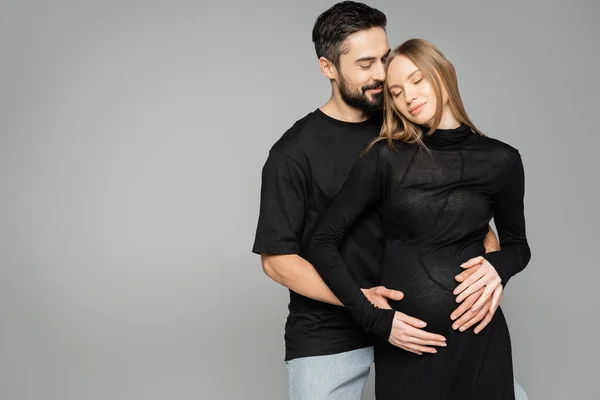 Positive bearded husband in black t-shirt hugging stylish pregnant wife in dress and closing eyes and standing isolated on grey, new beginnings and anticipation concept — Stock Photo