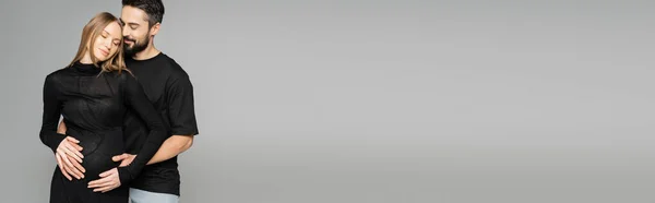 Hombre positivo y barbudo en camiseta negra abrazando a la mujer de pelo claro y embarazada mientras están de pie juntos aislados en gris, bandera, nuevos comienzos y concepto de anticipación - foto de stock