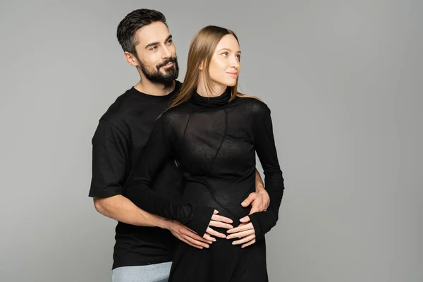 Улыбающийся и бородатый мужчина в черной футболке, обнимающий стильную беременную жену в платье и смотрящий вдаль изолированный от серого, новое начало и концепция предвкушения — стоковое фото