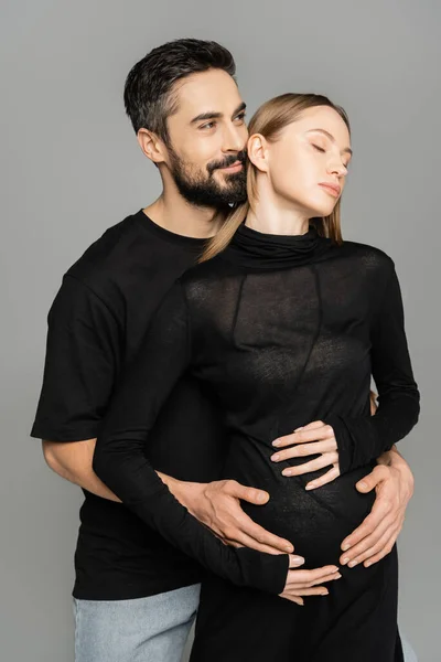 Улыбающийся и бородатый брюнетка мужчина в черной футболке обнимает светловолосую и беременную жену и смотрит в сторону изолирован от серого, новые начинания и концепции предвкушения — стоковое фото