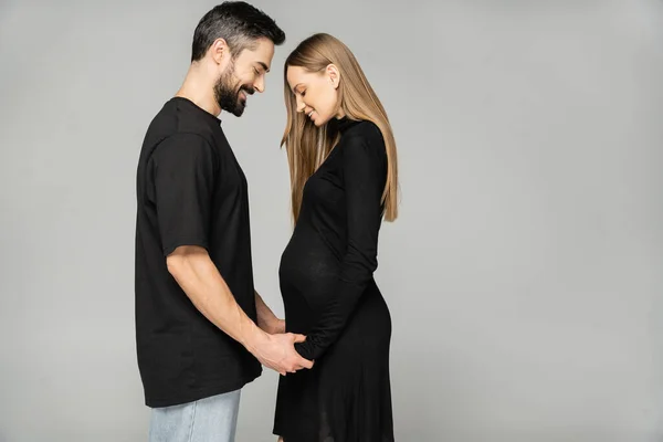 Side view of positive pregnant and stylish woman in black dress holding hands of bearded man while standing isolated on grey, new beginnings and anticipation concept, husband and wife — Stock Photo