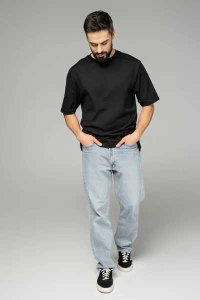 Longitud completa de hombre elegante y barbudo en camiseta negra y jeans tomados de la mano en bolsillos mientras camina sobre fondo gris, concepto de belleza masculina, confiado y carismático - foto de stock