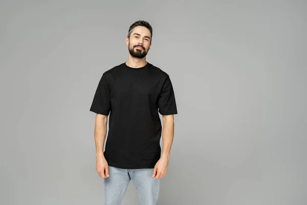Confident and relaxed bearded and brunette man in black t-shirt and jeans looking at camera while standing isolated on grey, masculine beauty concept, fashion-forward — Stock Photo
