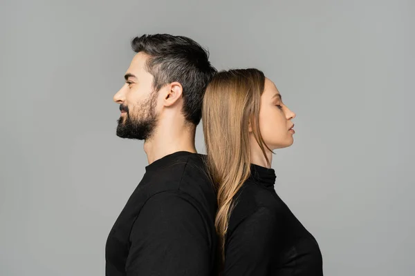 Seitenansicht einer entspannten und blonden Frau, die Rücken an Rücken mit einem bärtigen Ehemann in schwarzem T-Shirt steht, isoliert auf grau, Beziehungskonzept zwischen Mann und Frau — Stockfoto