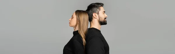 Side view of relaxed and bearded man in black t-shirt standing back to back with fair haired wife isolated on grey with copy space, husband and wife relationship concept, banner — Stock Photo