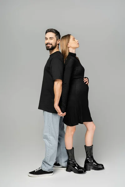 Full length of smiling bearded man looking at camera while holding hand and standing back to back with stylish pregnant wife in black dress on grey background, new beginnings and parenting concept — Stock Photo