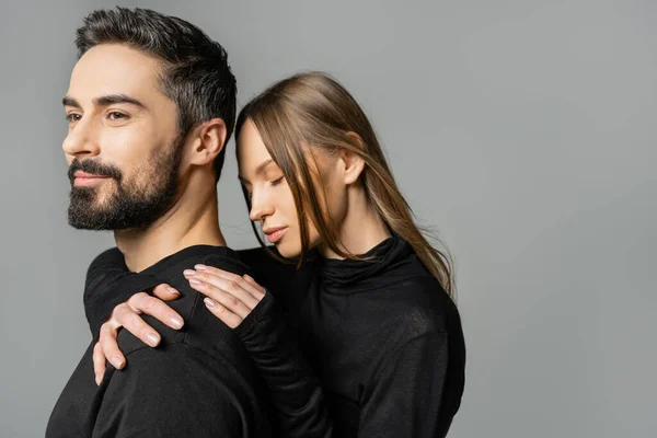 Fair haired woman in black clothes embracing smiling and bearded husband in t-shirt while standing together isolated on grey, husband and wife relationship concept — Stock Photo