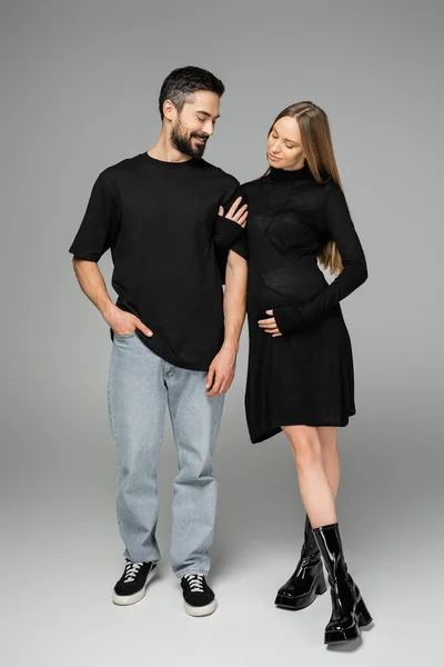 Full length of stylish pregnant woman in black dress touching arm of cheerful bearded husband while standing on grey background, new beginnings and parenting concept — Stock Photo