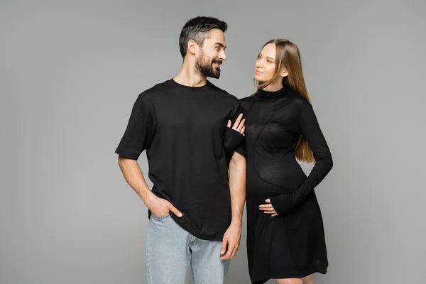 Положительная беременная женщина в черном платье, трогательная рука веселого бородатого мужа в футболке и джинсах, стоящего изолированно на сером, новом начале и концепции воспитания — стоковое фото