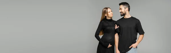 Stylish and pregnant woman in black dress touching arm of smiling bearded husband and standing together isolated on grey, new beginnings and parenting concept, banner — Stock Photo