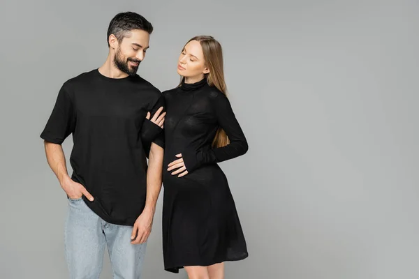 Стильная беременная женщина в черном платье трогает веселого мужчину в футболке и джинсах, стоя вместе изолированно на сером, новых начинаниях и концепции воспитания, мужа и жену — стоковое фото