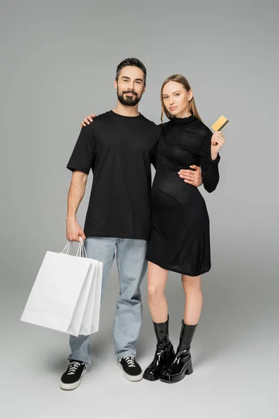 Стильна вагітна жінка в чорній сукні тримає кредитну картку і обіймає чоловіка з сумками, стоячи на сірому фоні, нові починання та концепція батьківства — стокове фото